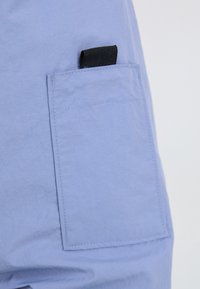 Pantalon en tissu bleu clair avec une poche latérale rectangulaire dotée d'une languette en tissu noir en haut.