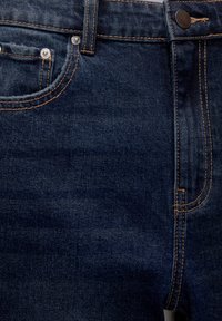 Jean en denim bleu foncé en gros plan montrant la poche avant, la petite poche à monnaie avec rivets, les passants de ceinture et la fermeture par bouton avec une couture contrastante orange.
