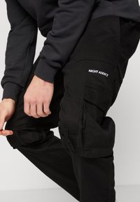 Černé cargo kalhoty s několika kapsami, nastavitelnými popruhy a bílým logem "NIGHT ADDICT" na stehně. Vyrobené z odolného materiálu.