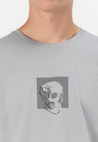 Šedé tričko s krátkym rukávom vyrobené z bavlny, s čiernobielou grafikou lebky obkreslenou v šedom štvorci. Hladká textúra.