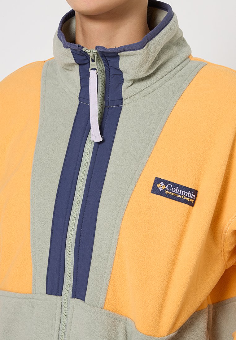 Persona con una chaqueta polar Columbia amarilla y gris, con un alto cuello y una cremallera central parcialmente abrochada.