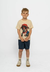 Beige t-shirt med en grafik av en hund i solglasögon och en röd keps, kombinerad med mörka shorts och beiga sneakers med ränder.