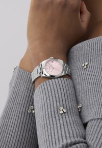 Montre en acier inoxydable argenté avec un cadran rond, un cadran rose et des accents argentés. Portée sur un poignet avec un pull gris à côtes.