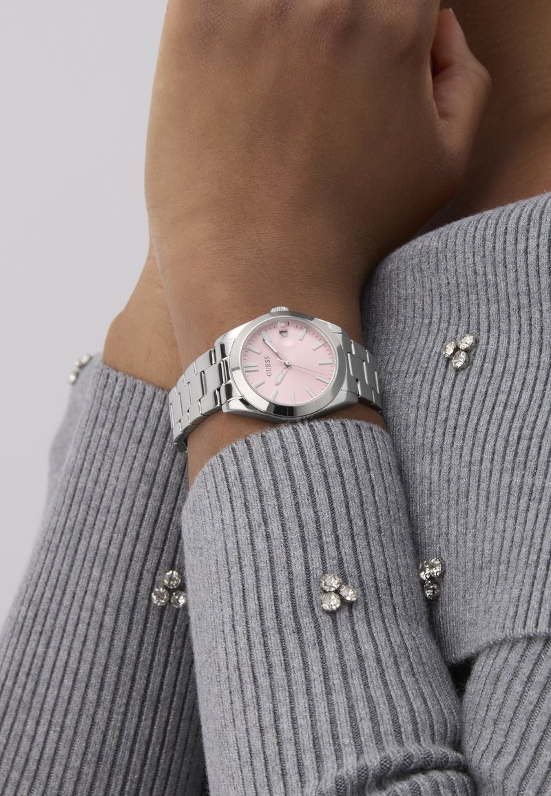 Montre en acier inoxydable argenté avec un cadran rond, un cadran rose et des accents argentés. Portée sur un poignet avec un pull gris à côtes.