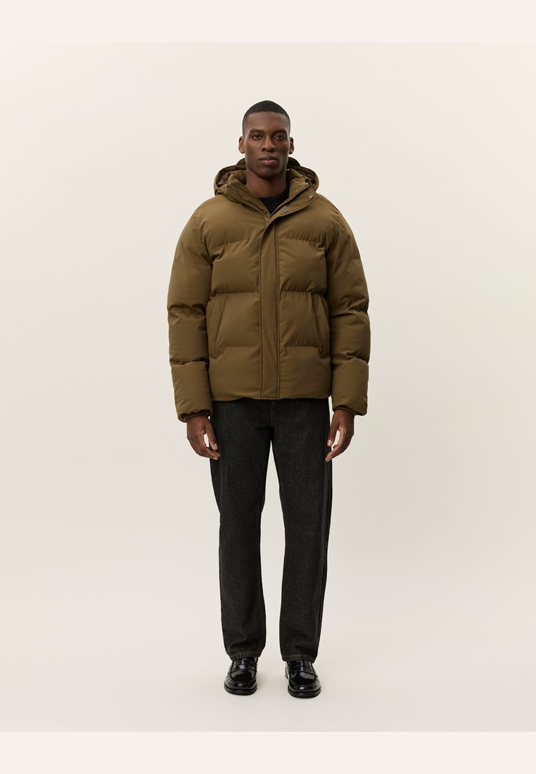 Chaqueta de abrigo marrón con capucha, que presenta un cierre frontal, diseño acolchado horizontal y material aislante. Combinada con pantalones oscuros y zapatos.