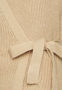 Beżowy sweter dzianinowy z teksturowanej tkaniny i detalem w postaci związania paska, w luźnym kroju z szerokimi rękawami.