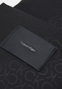 Melna auduma soma ar iegravētu CK rakstu un taisnstūra melnu ādas birku ar "Calvin Klein" sudraba burtiem.