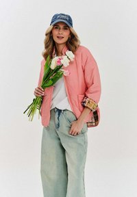 Růžová prošívaná bomber bunda s kostkovanou podšívkou, spárovaná s volnými džíny z světlého denimu. Modelka drží bílé a růžové tulipány a má na sobě modrou čepici.