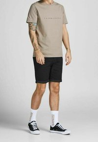 Beżowa koszulka z okrągłym dekoltem z czarnym logo, w zestawie z czarnymi szortami, białymi skarpetkami z tekstem marki oraz czarnymi sneakersami z białą podeszwą.