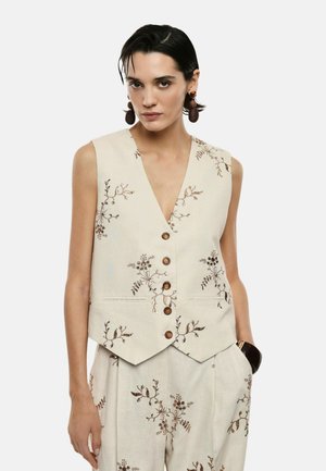 Donna con capelli corti e scuri indossa un gilet beige floreale senza maniche con bottoni e pantaloni abbinati, con grandi orecchini rotondi e braccialetto.