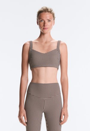 Femme aux cheveux blonds portant un crop top à carreaux marron et blanc et un pantalon assorti taille haute, debout sur un fond clair.