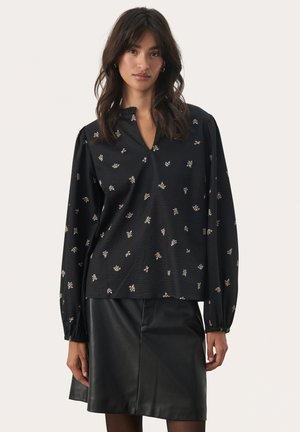Blouse noire avec un décolleté en V, des manches longues bouffantes et un tissu texturé avec de petites imprimés floraux dorés. Associée à une jupe en cuir noire.