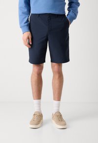 Marineblå shorts laget av lett stoff, med flat front, sidelommer og en avslappet passform. Brukt med beige sneakers og hvite sokker.