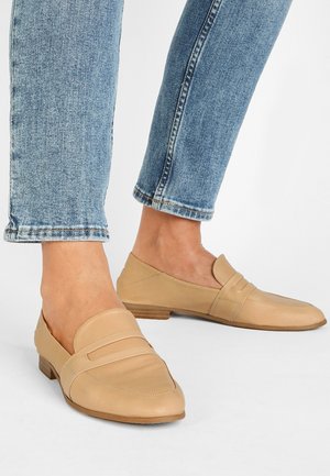 Mocassins en cuir beige à bout carré avec une bride décorative. Talon en bois bas, texture lisse et design minimaliste. Portés avec un jean bleu clair.