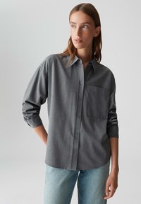 Woman wearing a loose gray button-up shirt with rolled-up sleeves and light blue jeans, looking off to the side against a plain background.