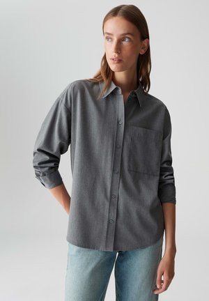Woman wearing a loose gray button-up shirt with rolled-up sleeves and light blue jeans, looking off to the side against a plain background.