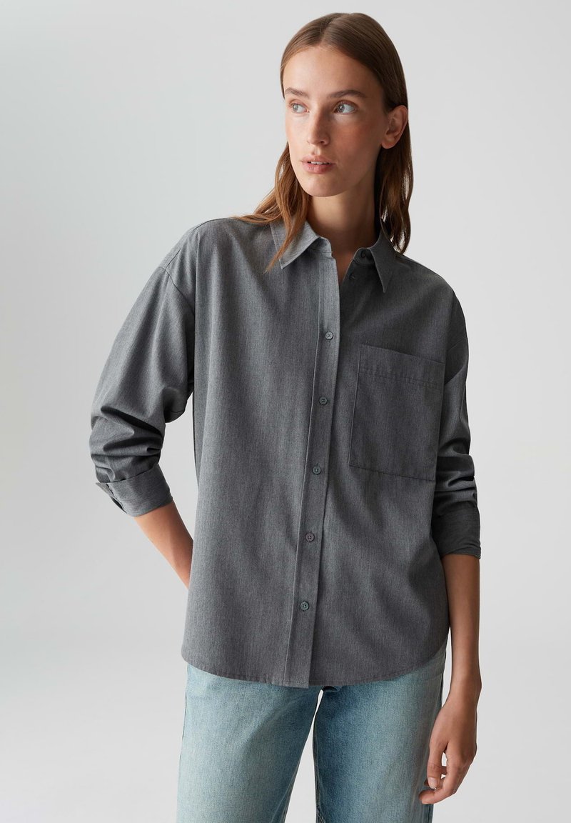 Woman wearing a loose gray button-up shirt with rolled-up sleeves and light blue jeans, looking off to the side against a plain background.