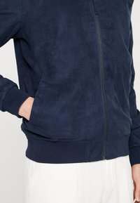 Námorně modrá zateplená kožená bunda na zip se bočními kapsami, žebrovanými manžetami a lemem. Hladký materiál s volným st�řihem, doplněná předním zipem.