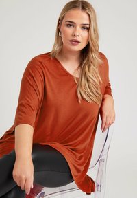 Rostfärgad, mjuk topp i tyg med V-ringning och trekvartslånga ärmar, kombinerad med svarta leggings i konstläder. Genomskinlig stol i bakgrunden.