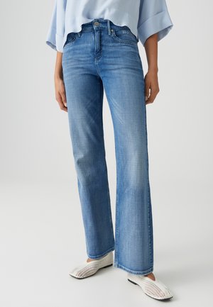 Vrouw die lichtblauwe jeans met wijde pijpen en witte geweven platte schoenen draagt, staand tegen een effen lichte achtergrond.