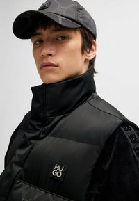 Black padded vest with a high collar, featuring a rubberized "HUGO" logo, and a textured fabric accent on the shoulder, worn with a matching cap.
