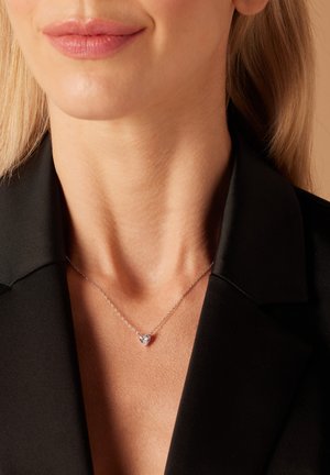 Zilveren ketting met een hartvormige hanger, gedragen op blote huid, met een fijne ketting en een gepolijste afwerking, op de achtergrond van een zwarte jas.