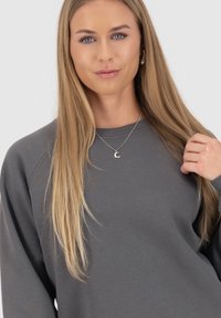Felpa grigia con una vestibilità rilassata, scollo rotondo e polsini a costine. Indossata con una delicata collana d'argento con un ciondolo a forma di luna crescente.