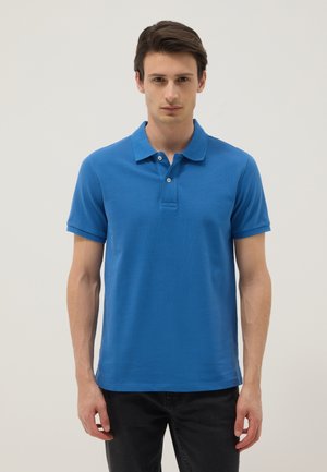 Junger Mann trägt ein hellblaues kurzärmeliges Poloshirt und schwarze Hose, steht vor einem einfarbigen hellen Hintergrund.