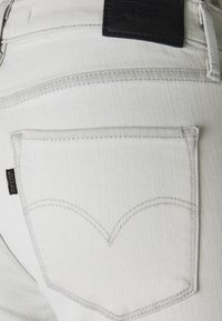Vaqueros de mezclilla blanca con un bolsillo trasero con un diseño de costura en forma de corazón, sutile costuras en gris y un parche de marca de cuero negro.