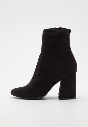 Czarne zamszowe botki za kostkę z ostro zakończonym noskiem, wysokim słupkowym obcasem i przeszyciami. Boczny zamek błyskawiczny ułatwiający zakładanie.