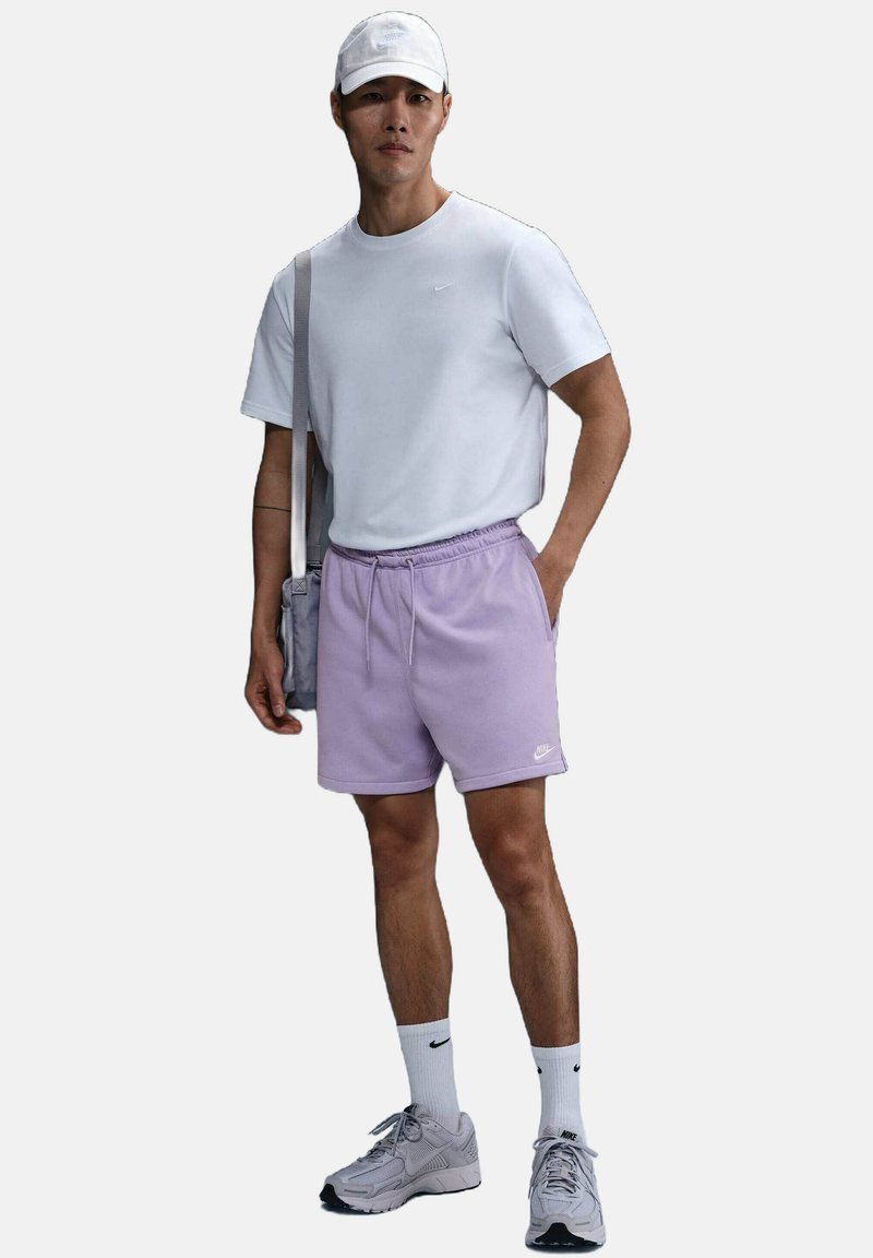 Camiseta blanca de manga corta de Nike, pantalones cortos morados claros con cinturilla, bolsa de hombro gris, calcetines blancos y zapatillas deportivas gris claro.