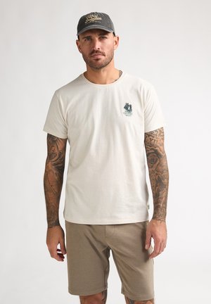 Man met mouwtattoos die een beige T-shirt, kaki korte broek en een donker petje draagt, staand tegen een effen lichte achtergrond.