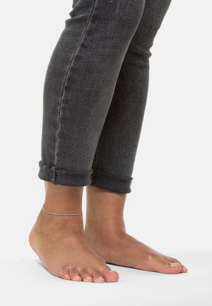Dunkelgraue Jeans aus Denim mit aufgerollten Säumen, die nackte Füße zeigen. Ein Handgelenk geschmückt mit einem zarten silbernen Kettenarmband, glatte Hauttextur.
