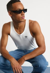 Bela rebrasta majica na naramenice z logotipom Adidas v črni barvi, v kombinaciji z modrimi kavbojkami. Model nosi črna pravokotna sončna očala in pozira v sproščenem položaju.