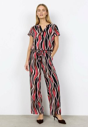 Vrouw draagt een bijpassend kort mouw topje en wijde broek met rode, roze, bruine en zwarte golvende strepen, staat met handen in de zakken.