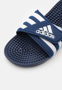 Blue slide sandal with three white stripes and an Adidas logo, featuring a textured sole with rounded dots for grip and comfort.