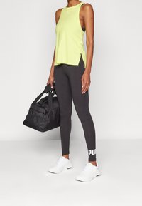 Světlé zelené tílko bez rukávů s postranními rozparky, v kombinaci s černými legínami s bílým logem "PUMA". Součástí jsou černá taška do posilovny a bílé tenisky.