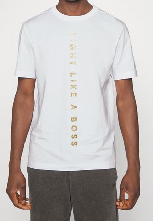Person trägt ein weißes T-Shirt mit vertikalem goldfarbenem Text "FIGHT LIKE A BOSS" und dunkelgraue Hose vor neutralem Hintergrund.
