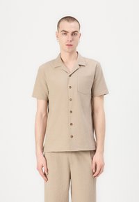 Camisa de manga corta beige con un acabado texturizado, cuenta con un bolsillo en el pecho y botones marrones, combinada con pantalones a juego.