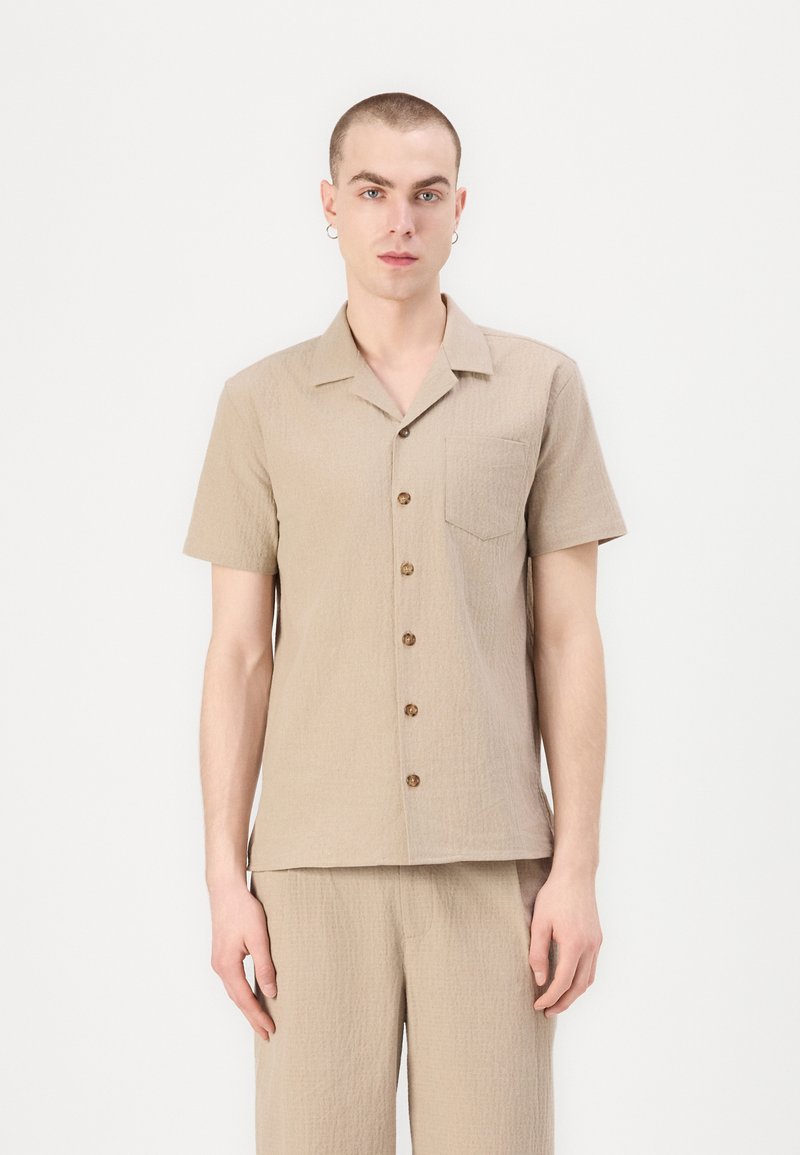 Camisa de manga corta beige con un acabado texturizado, cuenta con un bolsillo en el pecho y botones marrones, combinada con pantalones a juego.
