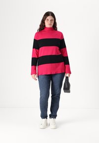 Long-sleeve, turtleneck top in bright pink with bold black stripes. Paired with blue denim jeans and white sneakers. Holding a black bag.
