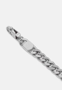 Silver chain bracelet with a textured link design and a lobster clasp. Features a rectangular charm with a logo detail.