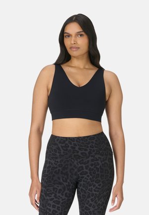 Vrouw met lang donker haar die een zwarte mouwloze crop top en zwarte legging met hoge taille en luipaardprint draagt, staand tegen een effen achtergrond.