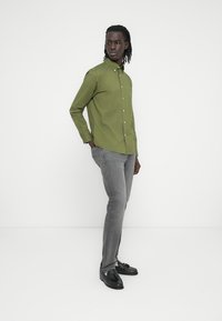 Πράσινο πουκάμισο με κουμπιά, μακριά μανίκια και γιακάς, σε συνδυασμό με γκρι τζιν στενής γραμμής και μαύρα παπούτσια, σε λευκό φόντο.