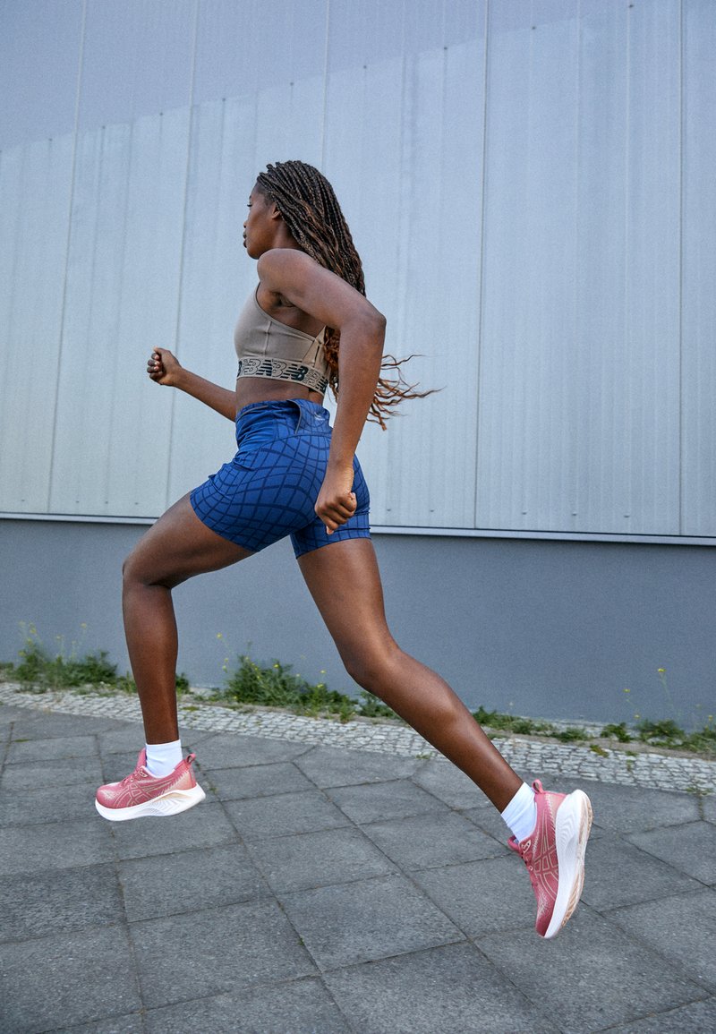 Sport-BH in Beige mit einem grafischen Bund, blaue gemusterte Shorts und pinke Laufschuhe mit weißen Sohlen, auf einem gepflasterten Außenweg.