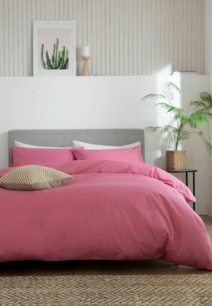 Doppelbett mit grauem Kopfteil und rosa Bettwäsche, beigem gemustertem Kissen, Topfpflanze auf dem Nachttisch und Kaktus-Kunst an weißer vertäfelter Wand.