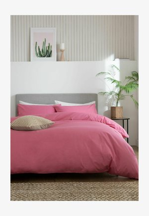 Doppelbett mit grauem Kopfteil und rosa Bettwäsche, beigem gemustertem Kissen, Topfpflanze auf dem Nachttisch und Kaktus-Kunst an weißer vertäfelter Wand.