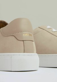 Beige semišové tenisky s bílými gumovými podešvemi, které mají zlatý akcent loga a prošívané detaily na patě a bocích.