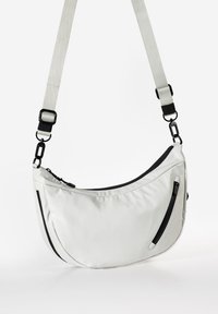 Weißer Schultertasche aus Stoff mit geschwungener Form, schwarzen Akzenten, einer Reißverschlusstasche und einem verstellbaren Gurt mit einem Metallclip.
