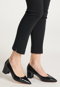 Zwarte leren pumps met hoge hakken en een puntige neus, voorzien van een blokhak en zij-snijdetails, gecombineerd met zwarte, taps toelopende broek.
